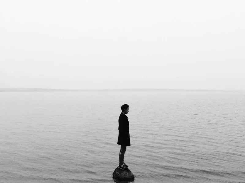 A man stands alone on a rock in the middle of an open body of water.
